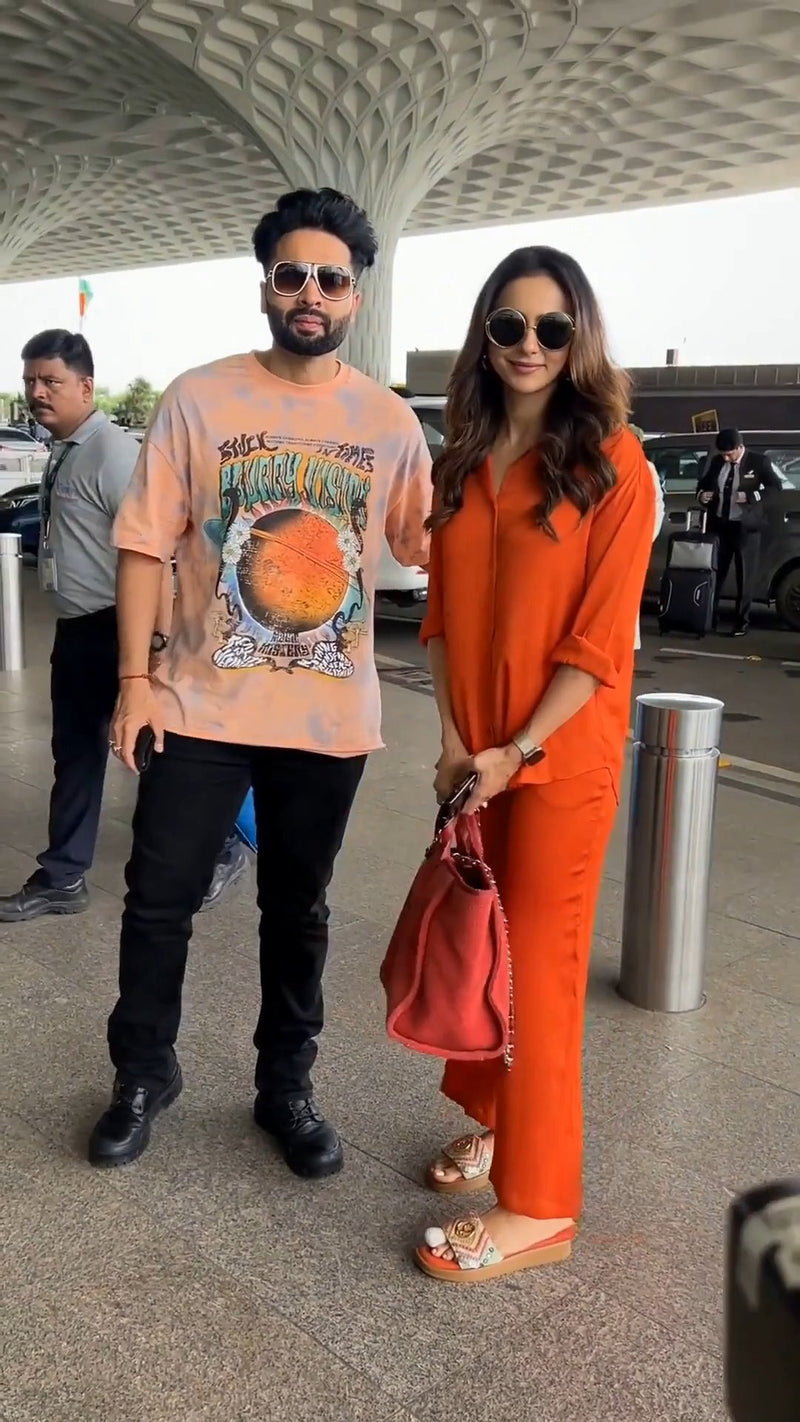 standing at an airport terminal holding a red handbag and her sunglasses in hand, wearing the solid orange co-ord set, beside a man in a graphic t-shirt near a pillar