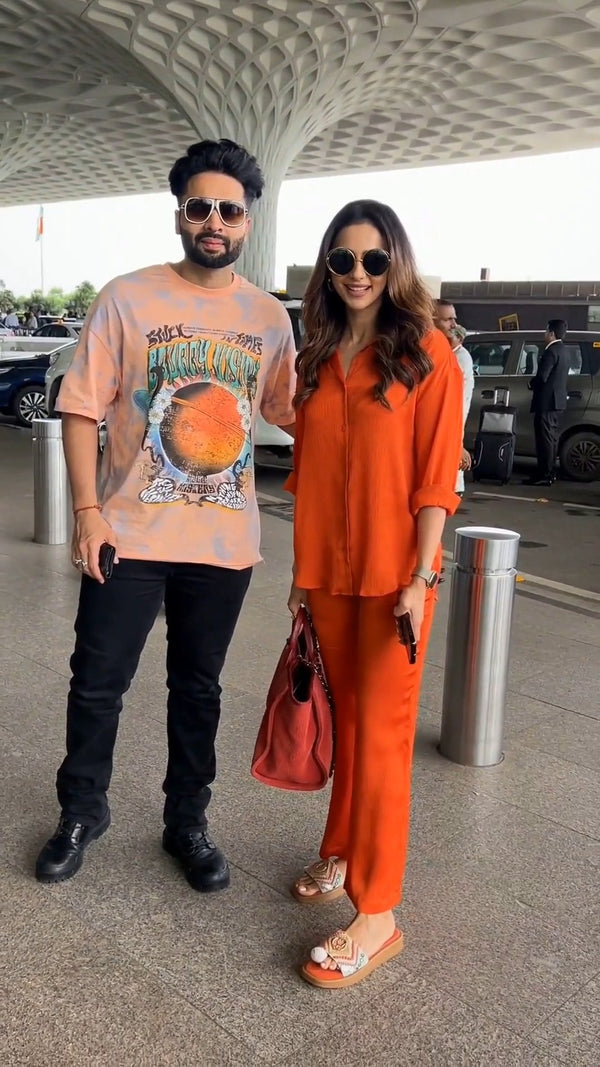 standing close beside a man at an outdoor airport pickup area with cars in the background, both wearing sunglasses, she in the solid orange co-ord set holding a red bag