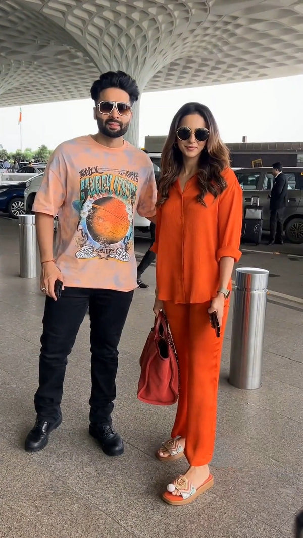 standing front-facing at an airport parking area in the solid orange co-ord shirt and pants, wearing sunglasses and holding a red handbag, beside a man in a graphic t-shirt