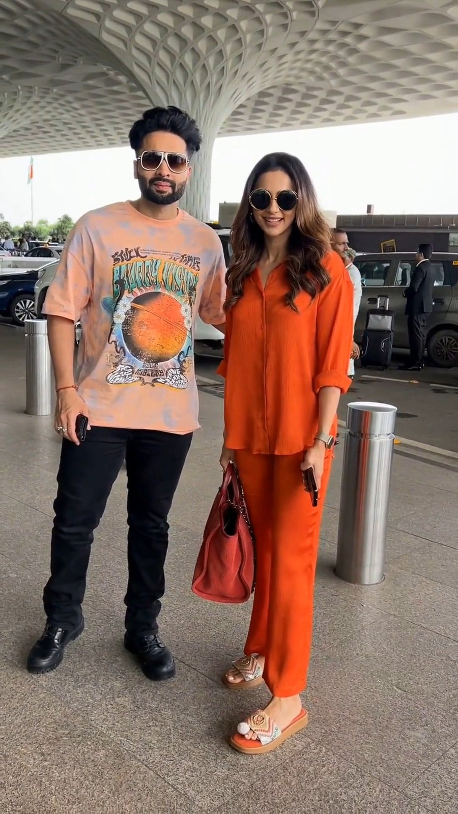 standing close beside a man at an outdoor airport pickup area with cars in the background, both wearing sunglasses, she in the solid orange co-ord set holding a red bag, photo 4