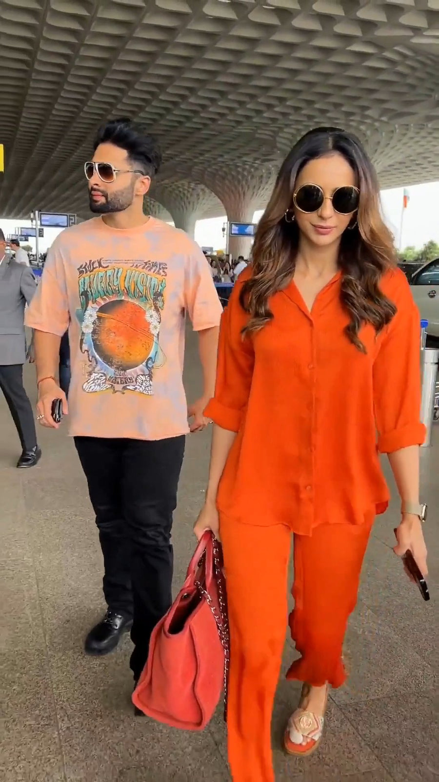 walking front-facing with a man under a covered airport walkway, wearing round sunglasses and the solid orange co-ord shirt and pants with rolled-up sleeves, holding a red handbag and her phone, photo 5