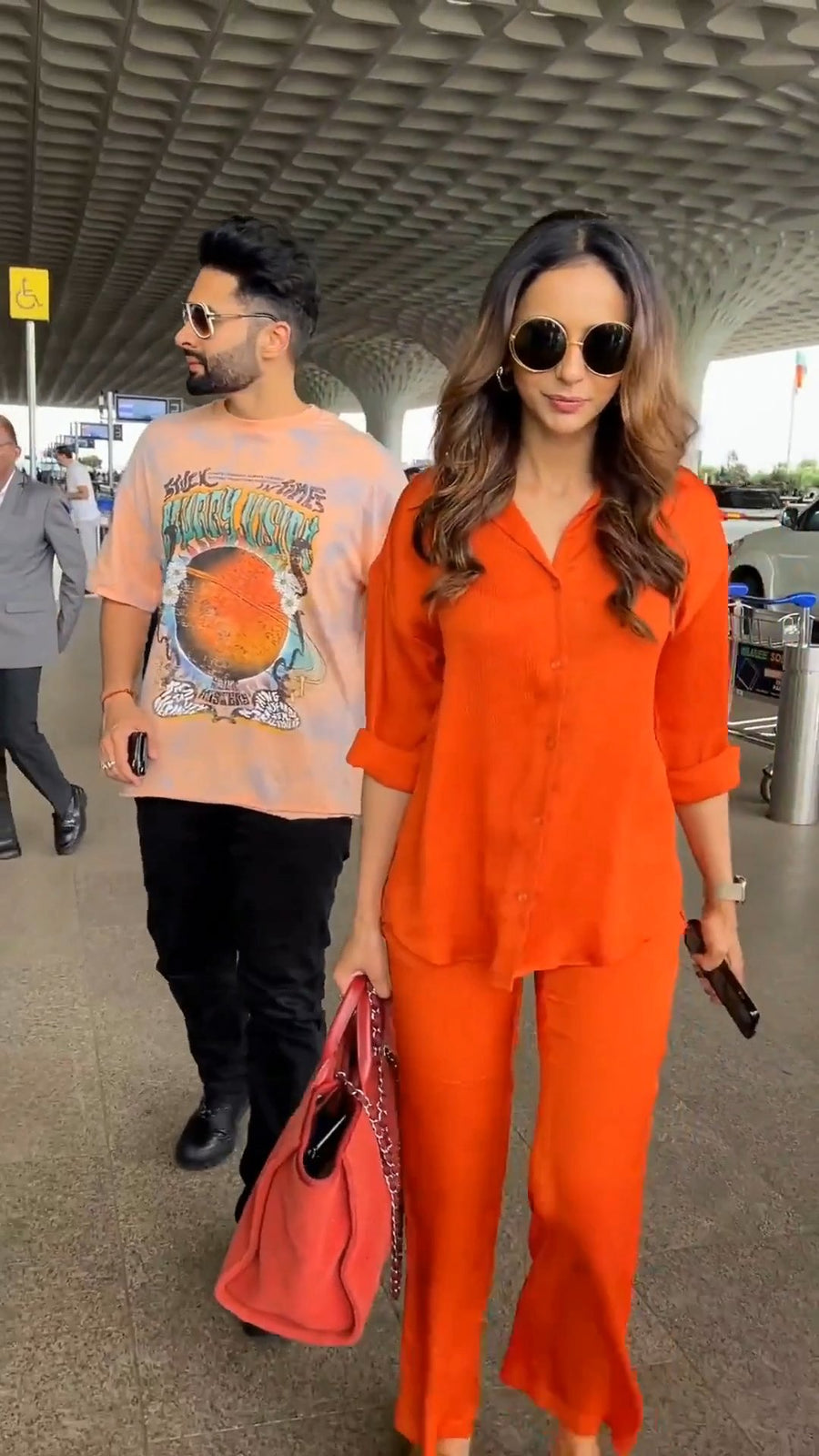 Close-up upper body shot of in round sunglasses at an airport terminal, wearing the solid orange co-ord shirt with rolled-up sleeves, holding her phone in hand, with a man in a graphic t-shirt beside her looking away, photo 6