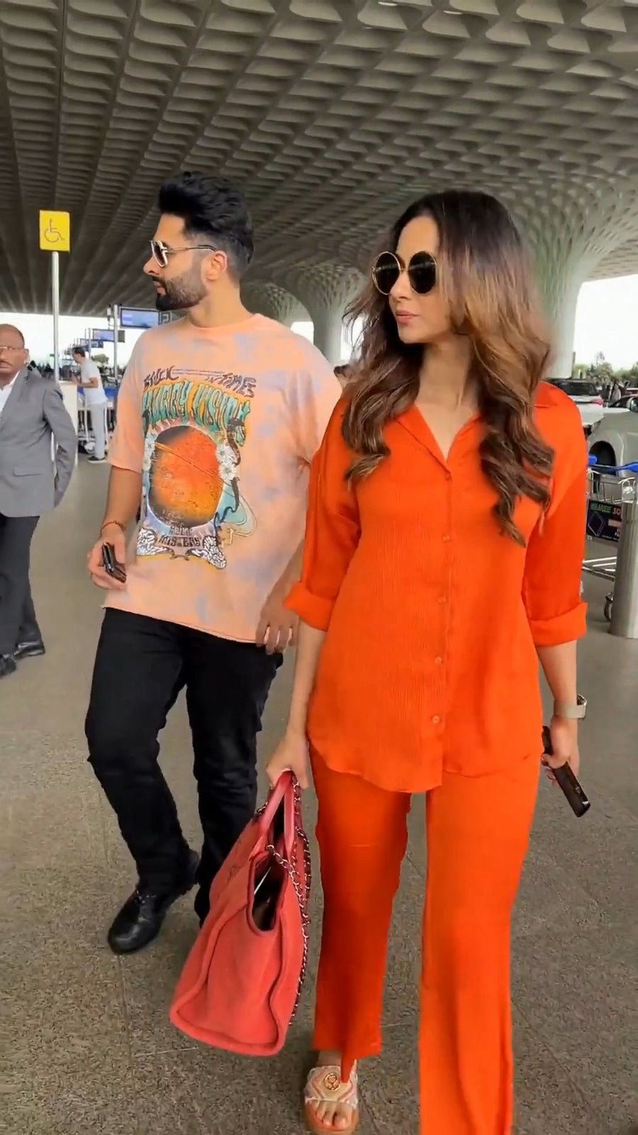 walking in side profile through an airport terminal in the solid orange co-ord set, holding sunglasses in her hand, with a man carrying a red bag beside her, photo 2