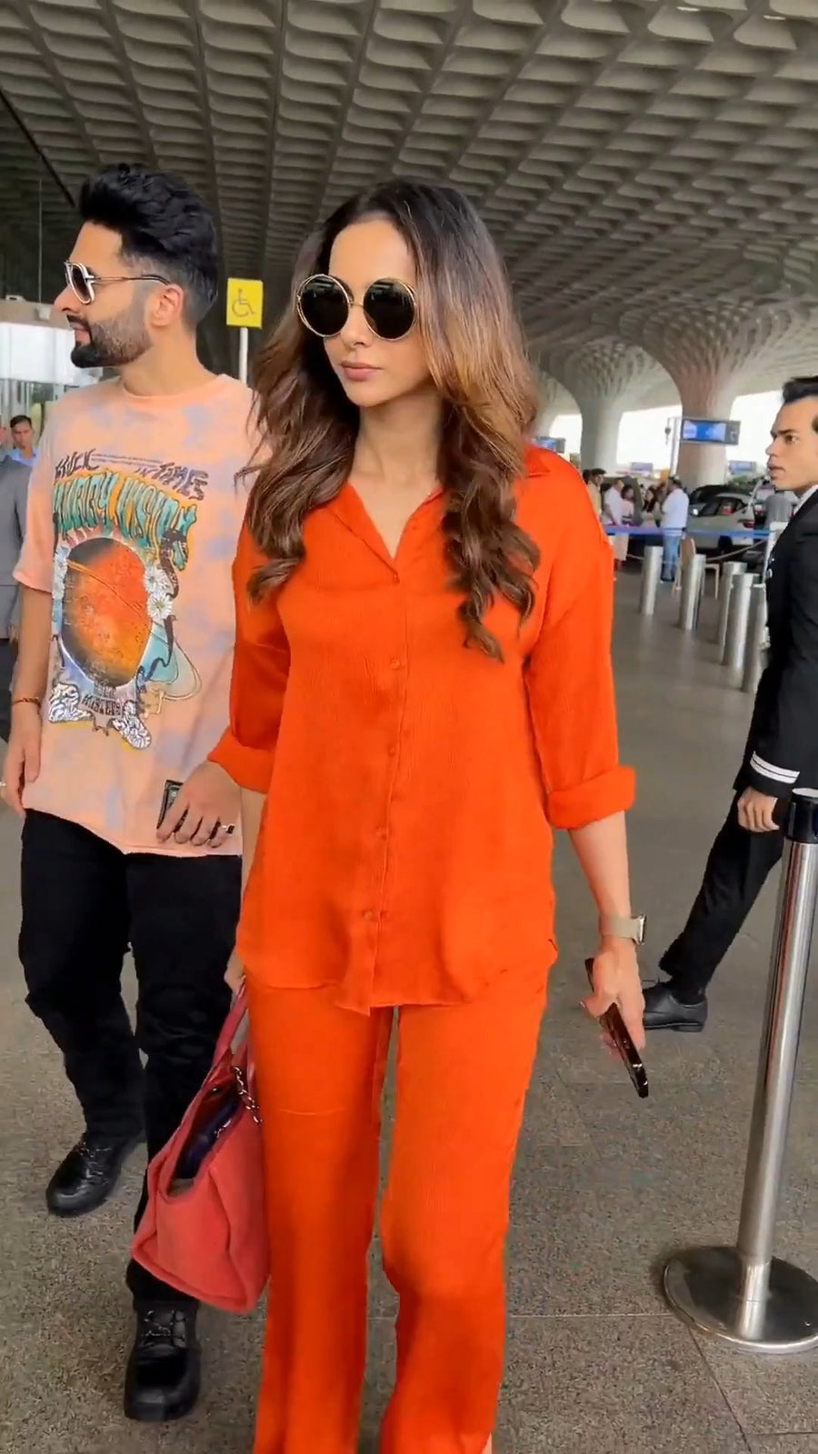 walking near an airport check-in barrier with a security guard visible in the background, wearing round sunglasses and the solid orange co-ord shirt and pants, holding her sunglasses case and a red handbag, beside a man in a graphic t-shirt, photo 7